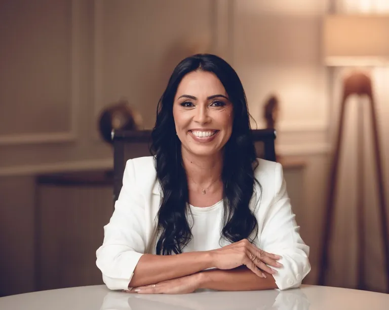 Geórgia Durães sorrindo, sentada à mesa, de blazer branco